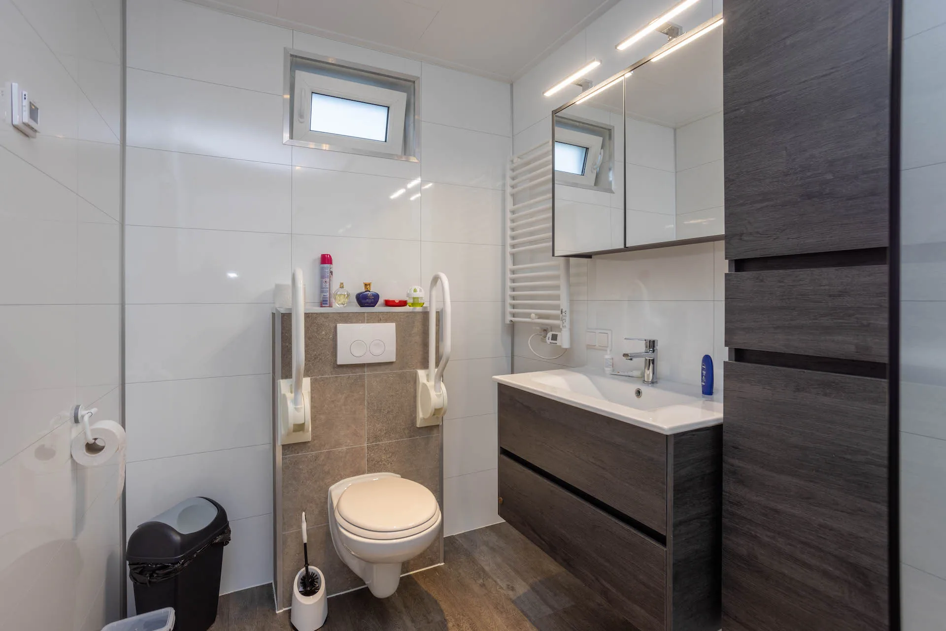 Badkamer met opklapbare wandbeugels rond het toilet en een houten vloer